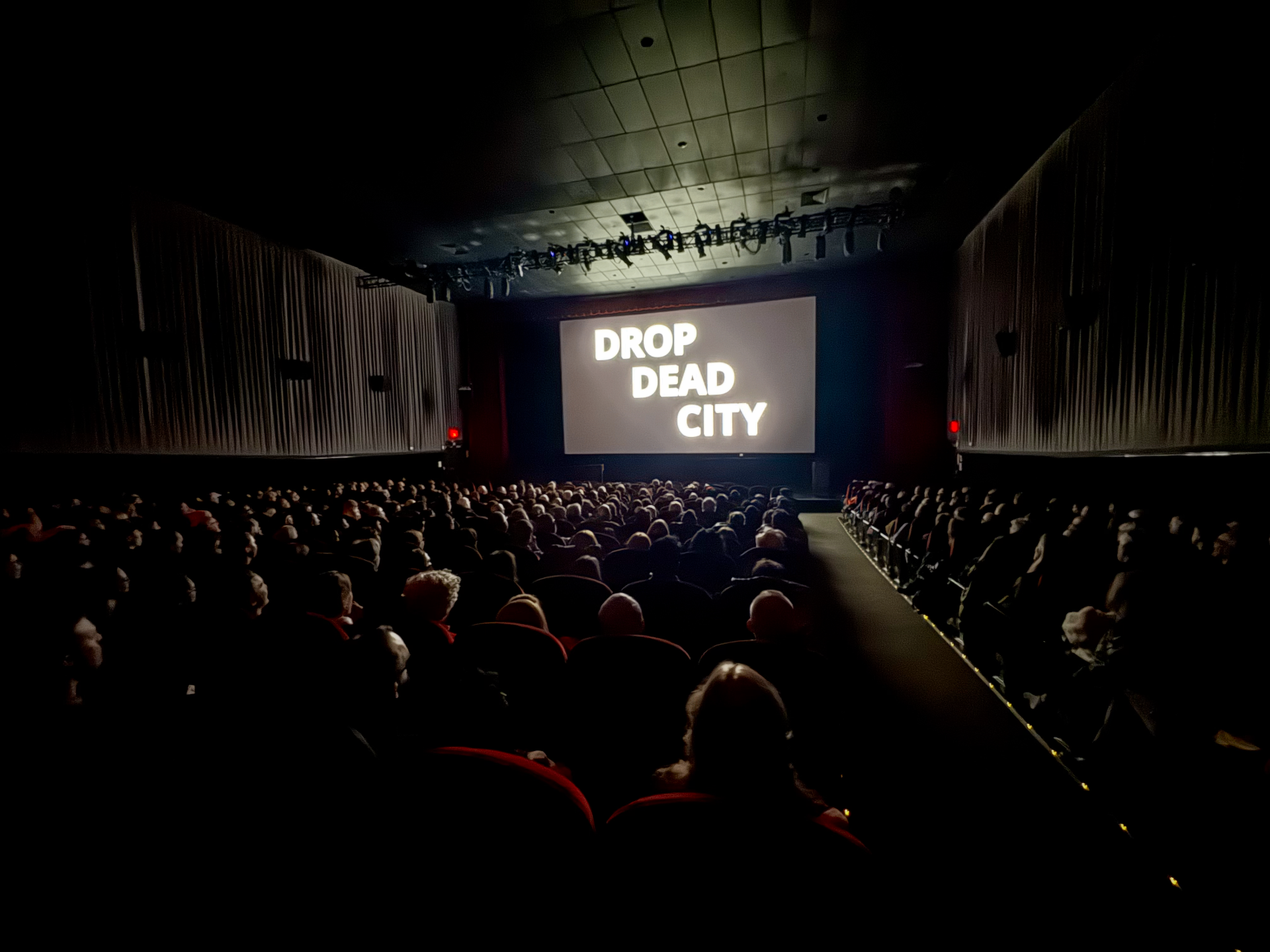 A sold-out theater audience watches the opening title card of Drop Dead City at a screening.